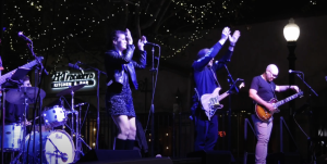 A live band performs on an outdoor stage at night under twinkling string lights. A female vocalist and a male guitarist are clapping with their hands raised, while another guitarist tunes his instrument at the SENSES Block Party.